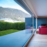 Casa sostenibile nel Canton Grigioni (CH)  - Una finestra che è molto più di un serramento. E lo sguardo inevitabilmente volge fuori, dove il verde del giardino e il blu del cielo contrastano con il rosso del divano ed il caldo colore del Rovere di Parkemo, Rovere, Collezione Versus Classic, spazzolato e oliato neutro, in formati misti.